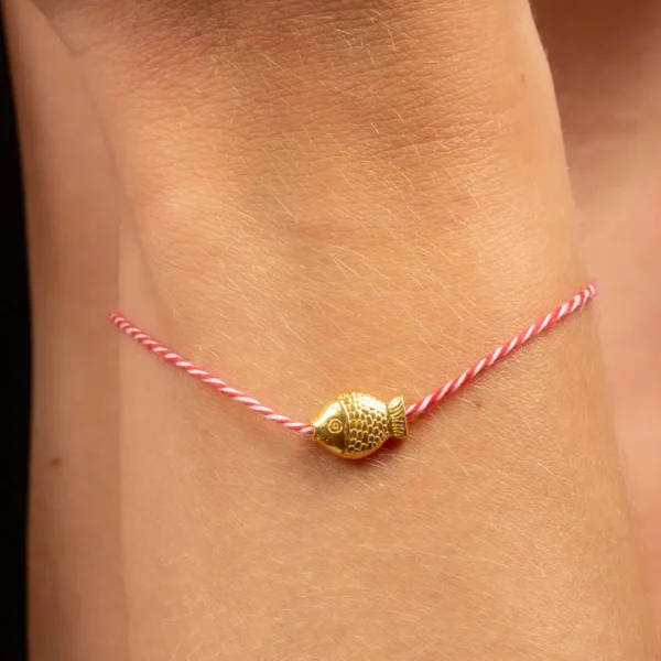 Gros plan sur le bracelet traditionnel grec, en fil torsadé blanc et rouge orné d’un poisson doré à l’or 24K, à nouer autour du poignet, du coffret. cadeau trois bijoux Eugénie, par Photyne