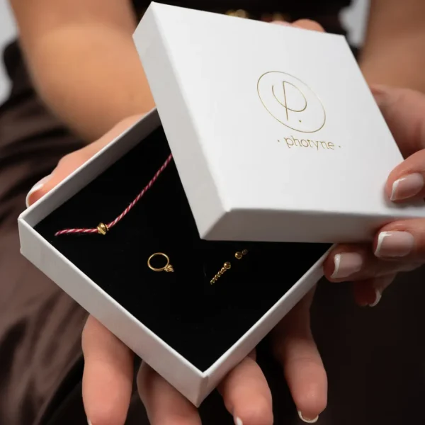 Coffret cadeau Photyne : boîte ouverte révélant un bracelet torsadé blanc et rouge orné d’une perle dorée, une paire de puces d’oreilles circulaires dorées et une bague fine torsadée dorée
