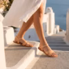 Sandales cuir pour femme à brides croisée couleur naturel , modèle Sitia de la marque Photyne -portées par une femme descendant des marches blanches en bord de mer, sous une robe blanche, dans une ambiance méditerranéenne ensoleillée.
