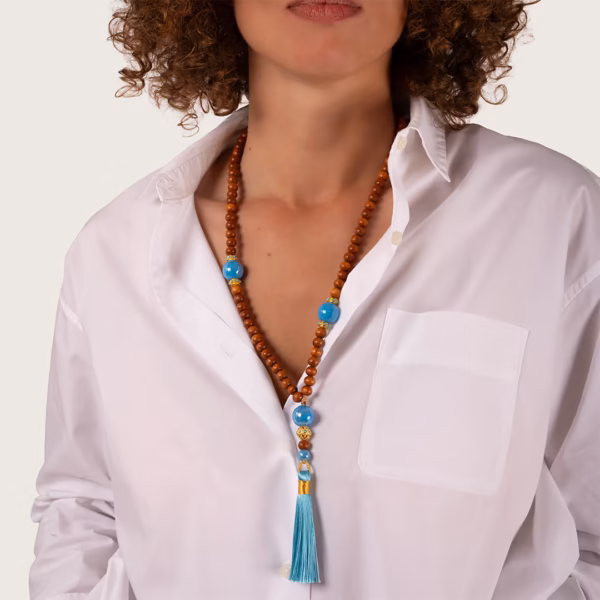 collier Nyx, perles de bois de bayong et de céramique bleue grecque - éléments dorés et pompon turquoise porté par une femme sur une chemise blanche entrouverte