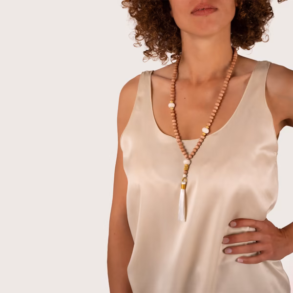Sautoir Chloe par photyne - perles bois et ornements dorés avec pompon blanc porté sur robe beige par une femme.